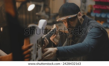 Close up of middle aged carpenters making electro guitar in stylish workshop. Professional tattooed artisan talks with colleague and tries on guitar fretboard. Handcraft and small business concept. Royalty-Free Stock Photo #2428432163