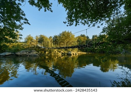 old rusty small wooden plank bridge over river in countryside, sunny summer day, green trees in background