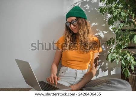 A stylish freelance woman with a laptop is sitting on the floor of a green ficus tree at home and working on an online project. A girl in a green hat and glasses works remotely from home, a modern web Royalty-Free Stock Photo #2428436455
