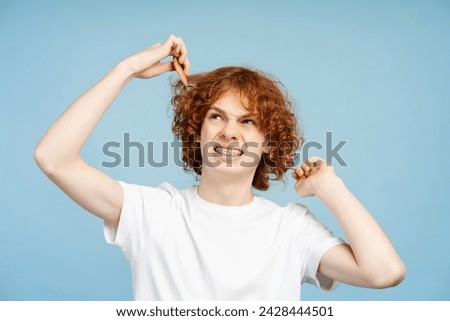 Sad curly haired ginger boy holding damaged curly hair, looking away isolated on blue background, studio shot. Hair care concept 