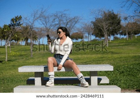 Young and beautiful Spanish brunette woman with sunglasses sitting on a park bench. The girl is casually dressed and consulting her mobile phone while enjoying the sunny spring day