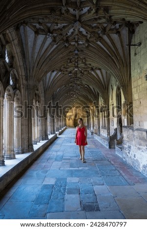 Young woman admires stunning arcades and tracery around that surround cloister. Tourist lady in red dress walks along a walkway in Canterbury Cathedral, known for its incredible Gothic architecture. Royalty-Free Stock Photo #2428470797