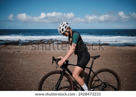Woman cyclist is using a bike computer during the training on Atlantic ocean background. Bike navigator. Cycling computer. Gravel road for cycling. Tenerife, Canary Islands, Spain.