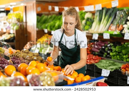 Fifteen-year-old girl who works part-time in a store as a trainee saleswoman puts tangerines on the counter for sale Royalty-Free Stock Photo #2428502953