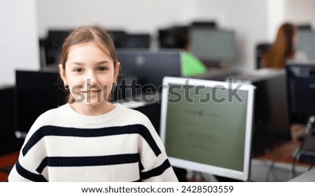 Positive interested teenage girl student standing in information technology classroom against background of rows of computers on desks Royalty-Free Stock Photo #2428503559
