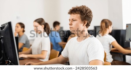 Focused teenager male student sitting at desk in college computer class, preparing for exam