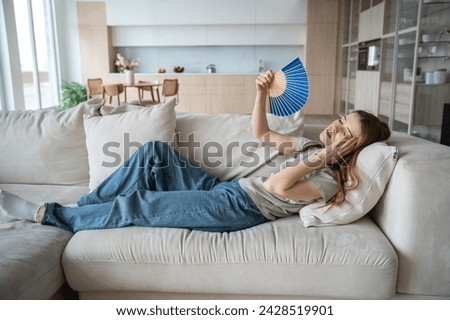 Unwell exhausted woman lying on home couch waving paper fan to cool down. Overheated female suffers from headache feeling malaise during stuffiness heatwave in apartment with broken air conditioning. Royalty-Free Stock Photo #2428519901