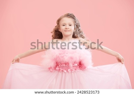 Children's holiday fashion. Beautiful little lady girl with lush curly hair and in a pink evening dress with feathers smiles. Pink studio background with copy space. Little Princess. Royalty-Free Stock Photo #2428523173