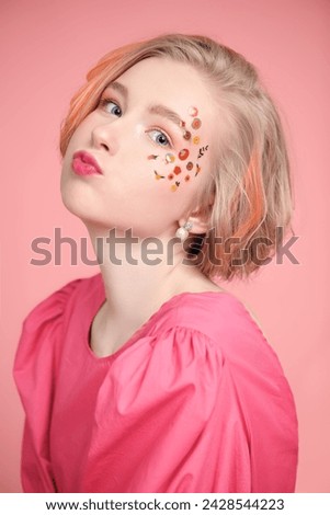 A cute blonde teenage girl with a short haircut poses in a pink dress with stickers in makeup and makes a funny face. Pink background. Spring-summer look.