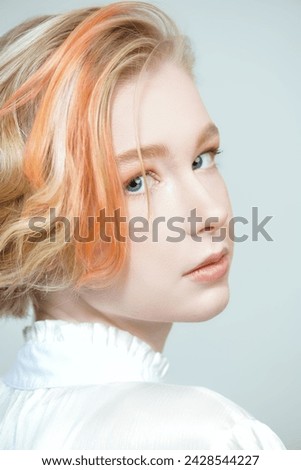 Adolescence. Beauty, style and fashion. A cute teenage girl with a short haircut and blonde hair with orange streaks poses in a white blouse on a white studio background.
