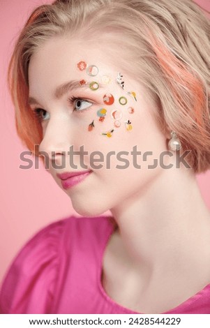 Spring-summer look. A cute blonde teenage girl with a short haircut poses in a pink dress with stickers in makeup and makes a funny face. Pink background.