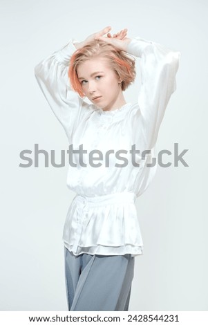 A cute teenage girl with a short haircut and blonde hair with orange streaks poses in a white blouse and grey trousers on a white studio background. Adolescence. Beauty, style and fashion.