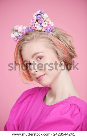 A cute blonde teenage girl with a short haircut poses happily in a pink dress and a lovely headband with floral kitty ears. Pink background. Kids and teenage fashion. Spring-summer look.