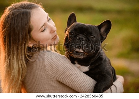 Cute young woman holding French bulldog, smiling at sunset while walking. Favorite dog in the arms of the owner in the park. Concept of pet care, adoption Royalty-Free Stock Photo #2428560485