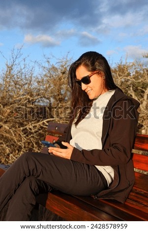 A young woman sitting on a bench, wearing sun glasses and looking at her cellphone in a blue day
