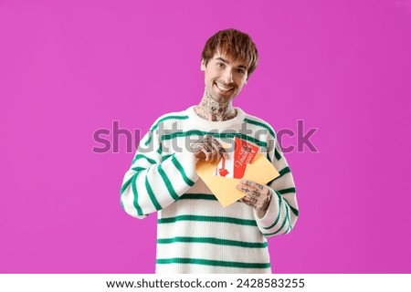 Young tattooed man with gift voucher in envelope on purple background