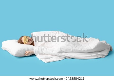 Beautiful young woman with soft blanket, pillow and mask sleeping on blue background