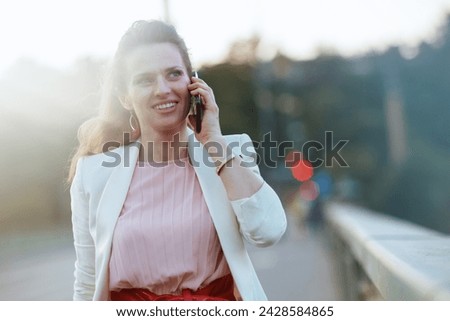 smiling modern female in pink dress and white jacket in the city using a smartphone on the bridge. Royalty-Free Stock Photo #2428584865