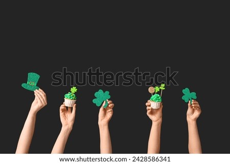 Female hands with tasty cupcakes and paper clovers for St. Patrick's Day on black background