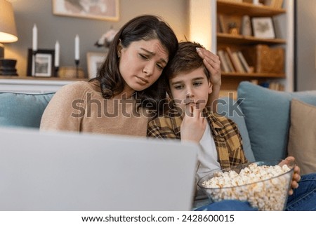 In their living room, a mother and her son share a cozy movie night. With popcorn in hand, they watch a sad film on the laptop, snuggled up on the sofa, finding comfort in each other's company. Royalty-Free Stock Photo #2428600015