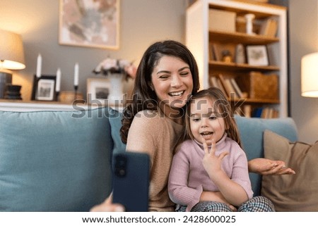 Smiling mother and daughter on video call using digital phone at home. while making video call and waving hello sitting on sofa. Woman and her cute little kid doing video conference with grandparents.