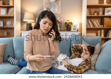 Mother measuring temperature of her ill kid. Sick child with high fever laying in bed and mother holding thermometer. Mother with cell phone calling to doctor