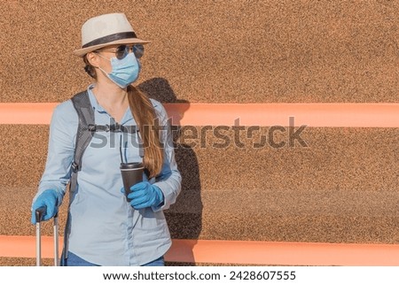 It is a view of young woman in blue medical mask and gloves with coffee cup near striped pink wall. It's sunny day. She is waiting a bus or a taxi.