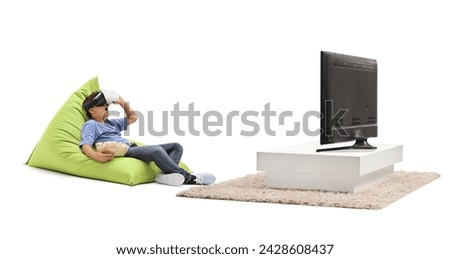 Kid with VR goggles eating popcorn and sitting on a beanbag in front of a tv isolated on white background Royalty-Free Stock Photo #2428608437