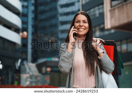 Charming pretty female standing in front of modern glass building. Successful corporate woman walking with shopping bags and talking on mobile phone while looking away with a smile.