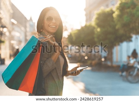 Attractive urban female looking at camera while standing on the street during the sunset holding colorful paper bags she got while shopping.