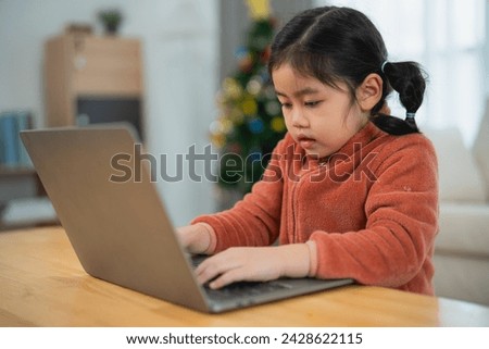 Asian Young Girl Focused on Using a Laptop at Home. A young girl with pigtails is engrossed in using a laptop on a wooden table, with a Christmas tree in the background.