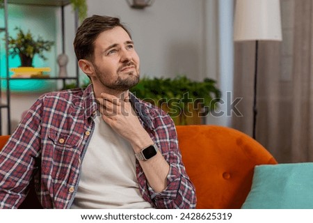 Upset Caucasian ill sick man holds hands over sore throat. Painful neck and frowning, thyroid disorders, suffering sore throat, tonsils inflammation. Young guy sitting on sofa at home room apartments Royalty-Free Stock Photo #2428625319