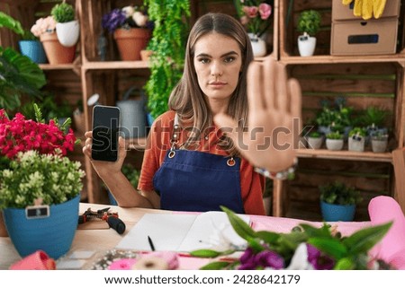 Hispanic young woman working at florist shop showing smartphone screen with open hand doing stop sign with serious and confident expression, defense gesture 