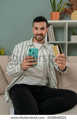 Vertical photo. Male sitting on the sofa is buying online with his smartphone and paying with the credit card with excited expression. Guy with mobile phone shopping in the living room using an app