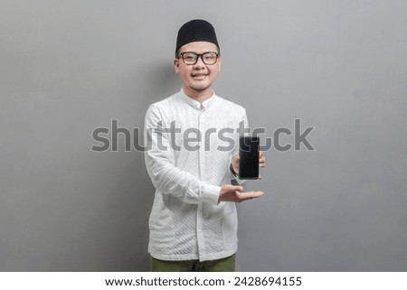 Portrait of an Asian Muslim man wearing a koko shirt and peci with shades of the fasting month, smiling showing his smartphone, isolated on a gray background