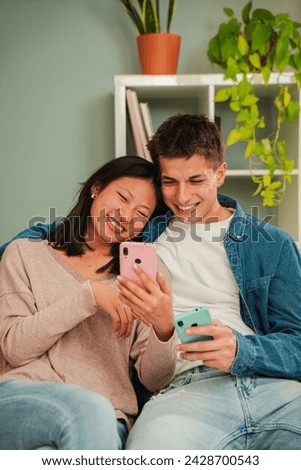 Vertical. Multiracial married couple using a cellphone to search apps sitting on living room sofa. Young people smiling and having fun together with a smart phone. Friends sharing on the social media