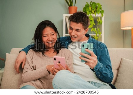 Real multiracial married couple using a cellphone to search apps sitting on living room sofa. Young people smiling and having fun together with a smart phone. Two friends sharing on the social media