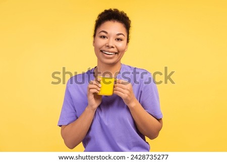 Human Emotions Concept. Photo of a delightful smiling young African American woman in a t-shirt, holding a mug of coffee, isolated on a yellow background. Morning energy theme Royalty-Free Stock Photo #2428735277