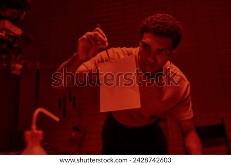 focused photographer holding tweezers with freshly developed photo paper in darkroom with red light