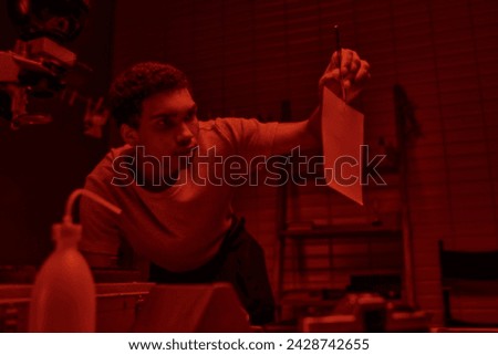 focused photographer holding tweezers with freshly developed photo paper in red light darkroom