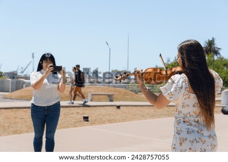 rear view of young latin woman busker violinist content creator playing violin outdoors while another girl films her for social networks.