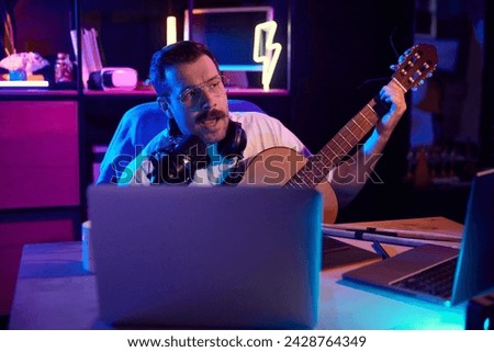 Artist composing music, vocalist playing on acoustic guitar in front of computer in neon light room, home office. Concept of youth people with social media and smart working, lifestyle, education.