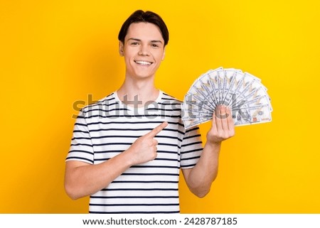 Portrait of pleasant student with stylish hairdo wear stylish t-shirt indicating at money in hand isolated on yellow color background