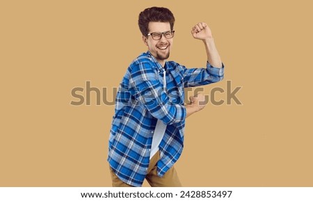 Funny, happy, nerdy student in casual clothes dancing to lively music at a party. Cheerful, joyful young man in a blue plaid shirt, light brown pants and glasses dancing isolated on a beige background