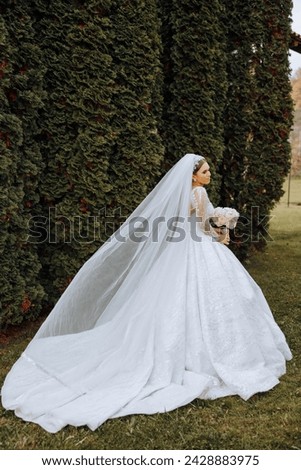 A brunette bride in a voluminous white dress holds a bouquet, poses. The veil is thrown into the air. Beautiful hair and makeup. Wedding photo session in nature. Long train of the dress