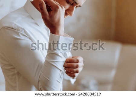 A man in a white shirt stands by the window in the room and fastens the buttons on his collar and sleeves. Watch on hand. Stylish business portrait of a man, close-up photo. The groom is preparing. Royalty-Free Stock Photo #2428884519
