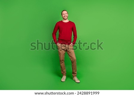 Full size photo of pleasant satisfied man dressed red pullover brown pants holding arms in pockets isolated on green color background