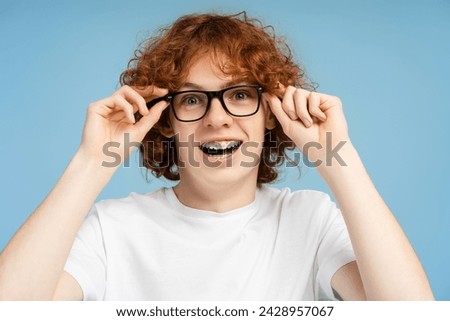 Headshot of a nerdy, curly red haired teenage boy with braces, equipped with eyeglasses, looking directly at the camera with his mouth wide, isolated on a blue background