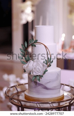 Elegant, three-tiered white wedding cake with fresh flowers on a golden stand. Backdrop of golden chains against greenery. Concept for wedding planners showcasing luxurious desserts. Royalty-Free Stock Photo #2428977707