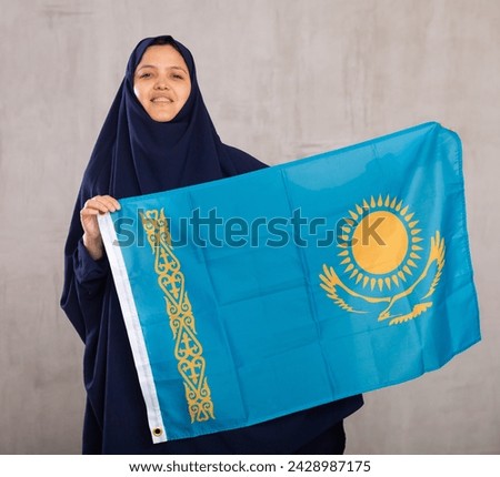 Portrait of young smiling woman in hijab posing with large flag of Kazakhstan in studio
